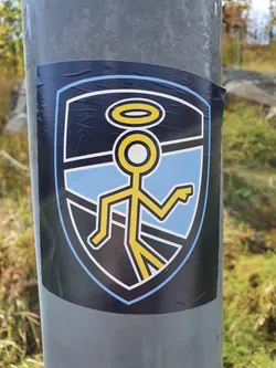 Street sticker A sticker featuring a minimalist design of a yellow stick figure with a halo, walking on a path depicted inside a shield shape. The style is simple and graphic, with a light blue and dark background.