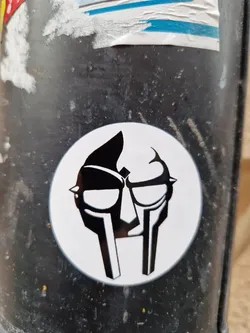 A round sticker featuring the iconic MF DOOM mask. The design is simple yet striking, with the mask rendered in black and white against a white background. This sticker is a great example of fan art and a tribute to the late rapper.