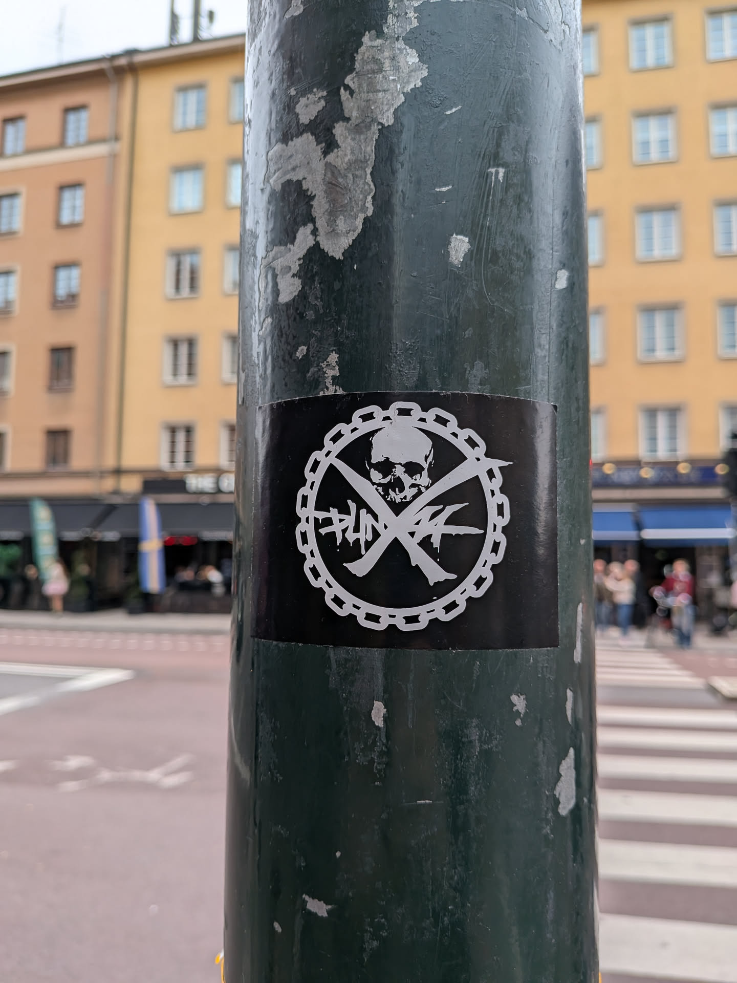 A rectangular black sticker affixed to a dark green metal pole. The sticker features a white circular graphic in the center. The outer ring of the circle is depicted as a chain. Inside the circle, a human skull sits atop two crossed bones that form an 'X'. Stylized, graffiti-like text is written across the bones, reading 'PUNK' on the left and 'AF' on the right.