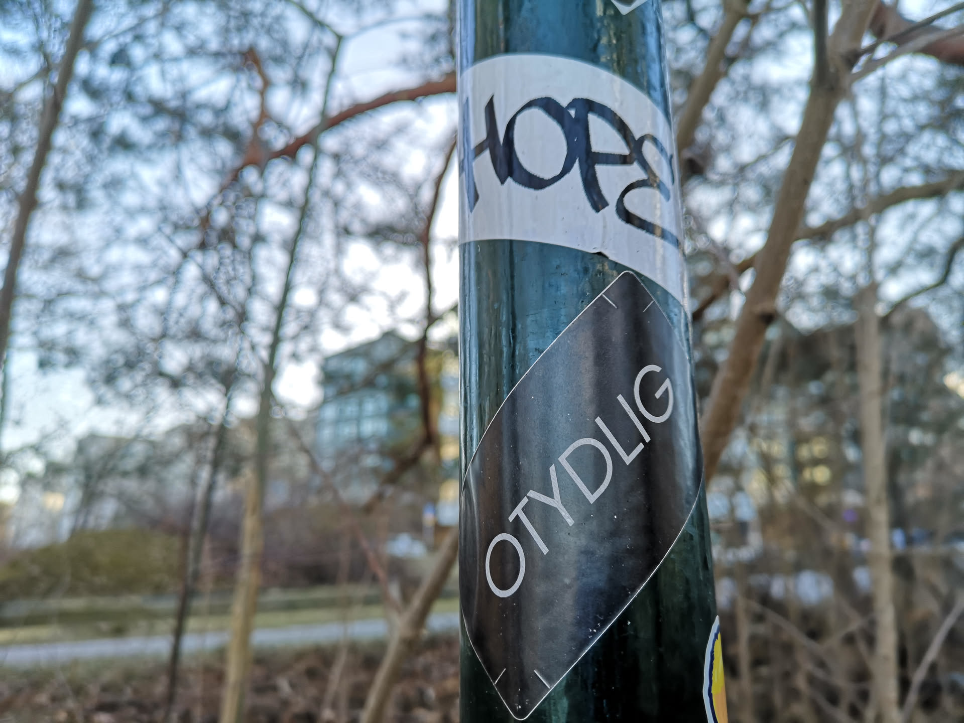 The central sticker is a black, rectangular shape, oriented diagonally on a dark green pole. It features the word "OTYDLIG" in clear, sans-serif white capital letters, running from the bottom left to the top right. The sticker has a subtle white outline or border, and its surface shows minor scuffs and dust, indicating it's an outdoor application. The background of the sticker is a solid, matte black.