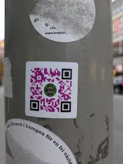 A sticker with a QR code in the center.  The QR code has a small logo in the middle and purple accents. The text around the bottom of the sticker says "Gå med i kampen för en fri värld" which translates from Swedish to "Join the fight for a free world".  There's another sticker above it with partial text visible: "www.knappt"