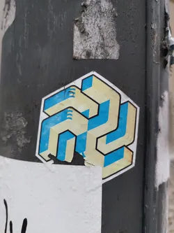Street sticker Close-up view of a sticker featuring an abstract design. The design is primarily composed of geometric shapes in beige and teal, creating a three-dimensional effect. The sticker is affixed to a dark grey pole, and a portion of the sticker is slightly damaged or peeled.