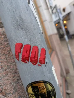 Street sticker The sticker is rectangular with rounded corners, featuring the word 'FAAA' in a prominent, bold, sans-serif typeface. The letters are a vibrant red color, with a slightly distressed or textured appearance, giving them a somewhat grungy or worn look. There are visible water droplets on the clear plastic overlay of the sticker, indicating it's exposed to the elements. The sticker is adhered to a grey, textured metal pole, with some clear adhesive visible around the edges of the red lettering. The background of the sticker itself is transparent, allowing the grey pole to show through where there is no red ink.