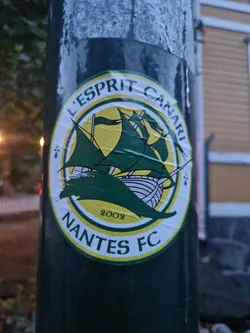 Street sticker A green and yellow circular sticker featuring the Nantes FC logo. The logo includes a ship, the text "L'Esprit Canari" and "Nantes FC", and the year 2002. The sticker is affixed to a dark green pole.