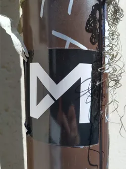 A black and white sticker with a stylized letter M.  The design is geometric and modern. The sticker is affixed to a brown pole.