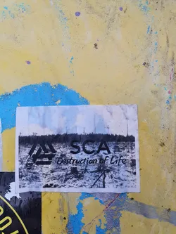 Street sticker A black and white photo of deforestation with the letters SCA and the text Destruction of Life printed on it. The sticker is placed on a yellow painted wall.