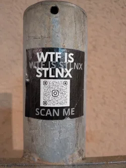 A rectangular sticker with a black background adhered to a textured grey metal pole. The top portion features white, bold, sans-serif text that reads "WTF IS" on the first line, followed by "WTF IS STLN X" on the second line, with "STLN X" being more prominent. Below the text, there is a large white QR code with a black Instagram camera logo at its center. At the very bottom, in smaller white text, it instructs "SCAN ME". The sticker shows slight creasing and wear, indicating it's a physical sticker on an outdoor surface.