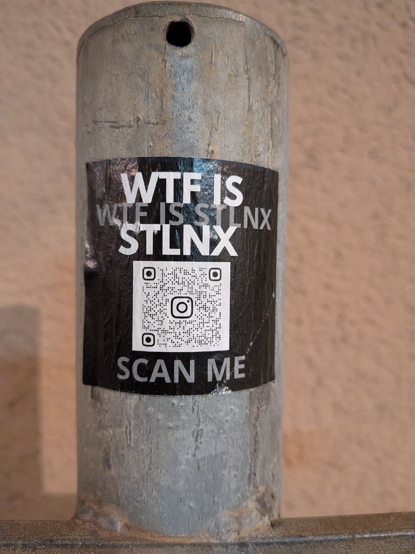 A rectangular sticker with a black background adhered to a textured grey metal pole. The top portion features white, bold, sans-serif text that reads "WTF IS" on the first line, followed by "WTF IS STLN X" on the second line, with "STLN X" being more prominent. Below the text, there is a large white QR code with a black Instagram camera logo at its center. At the very bottom, in smaller white text, it instructs "SCAN ME". The sticker shows slight creasing and wear, indicating it's a physical sticker on an outdoor surface.