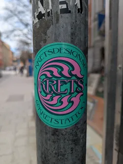 Street sticker This round sticker features a turquoise background with stylized pink waves and the artist's name 'KRETS' in the center. The artist's website 'KRETSDESIGN.SE' is printed along the top edge, and their Instagram handle '@KRETSTATTOO' is at the bottom. The sticker is affixed to a weathered pole.