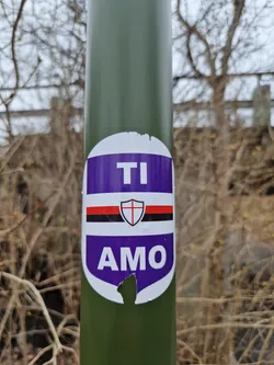 A purple and white oval sticker with the words "TI AMO" in white lettering and a shield emblem is affixed to a green pole. The sticker appears to be slightly damaged, with a small tear near the bottom. The background of the image shows bare branches and undergrowth suggesting an outdoor location. 