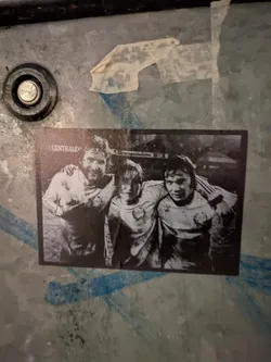 Street sticker A black and white sticker featuring a photograph of three men, likely footballers, posing with their arms around each other. They are wearing jerseys with some sort of emblem and what appear to be Adidas stripes on the sleeves. The background suggests an outdoor setting, possibly a sports field, and the image has a vintage feel. The sticker is slightly peeling at the top right corner and appears to be adhered to a textured, metallic surface with some blue markings.