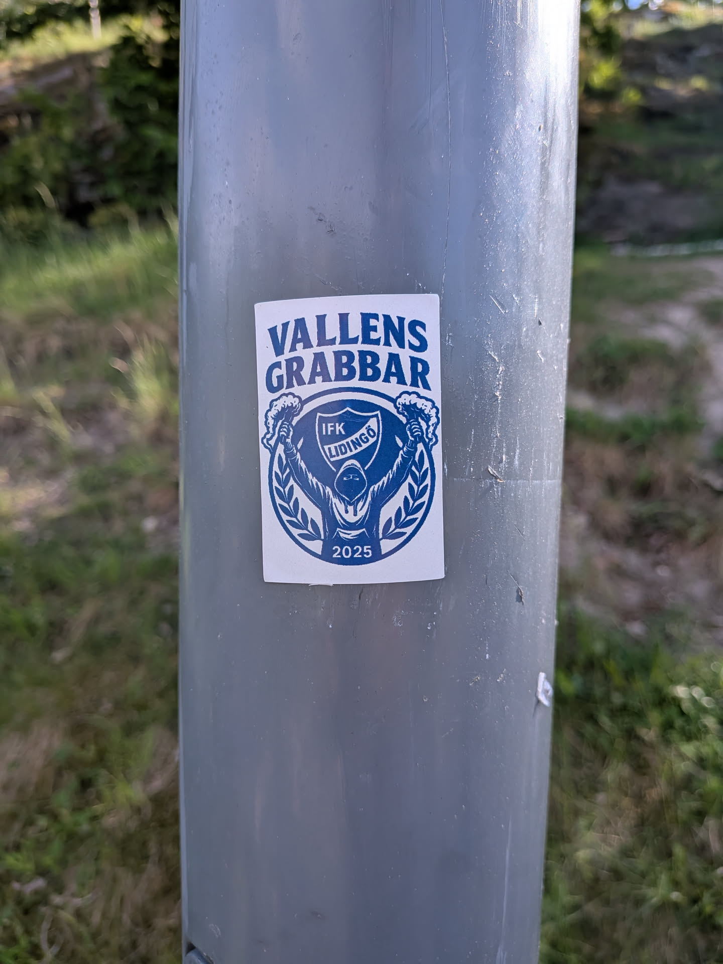 A rectangular sticker, predominantly white with bold blue graphics, affixed to a grey pole. The top features the arched text "VALLENS GRABBAR" in a strong, sans-serif font. Below this, a circular emblem is framed by stylized laurel branches. Inside the circle, a hooded figure is depicted from the waist up, with arms raised, holding what appears to be a flaming torch or flare in their right hand. Over the figure's chest, a shield contains the text "IFK LIDINGÖ". At the very bottom of the circular design, the year "2025" is inscribed. The overall aesthetic is graphic and robust, indicative of a sports fan or supporter group.