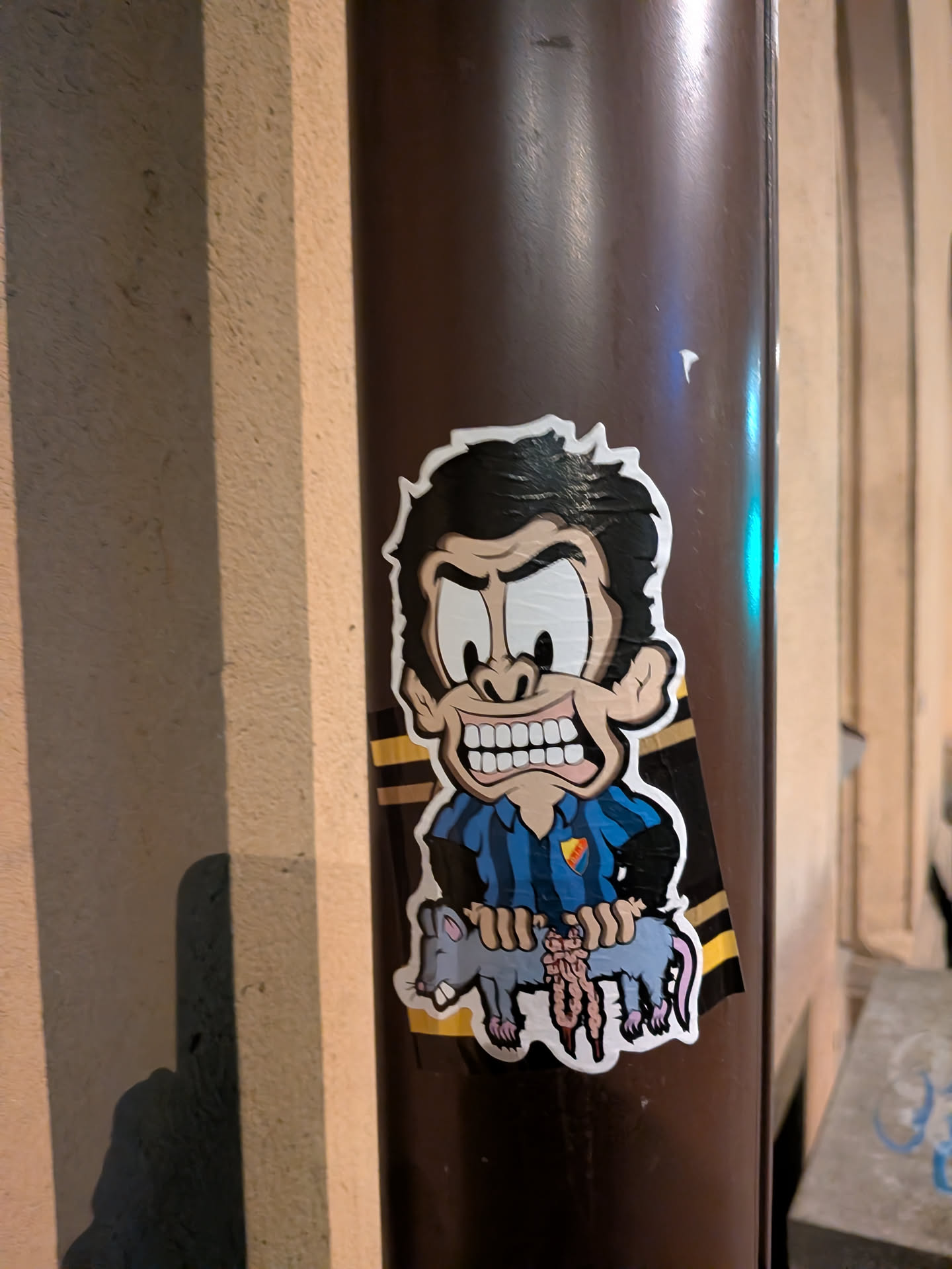 A cartoon sticker featuring an angry-looking man with dark, messy hair, wide eyes, furrowed brows, and a grimacing expression showing many white teeth. He wears a blue and black striped shirt, resembling a football jersey, with a yellow shield-shaped crest containing a red symbol on the left chest. The character is holding a grey rat by its front and back legs, which appears to be disemboweled with pinkish-red intestines spilling out. Behind the character, diagonal yellow and black stripes are visible, strongly suggesting a connection to AIK Fotboll, whose fans are often called 'Gnagare' (rodents). The sticker has a distinct white outline and is affixed to a dark brown pole.