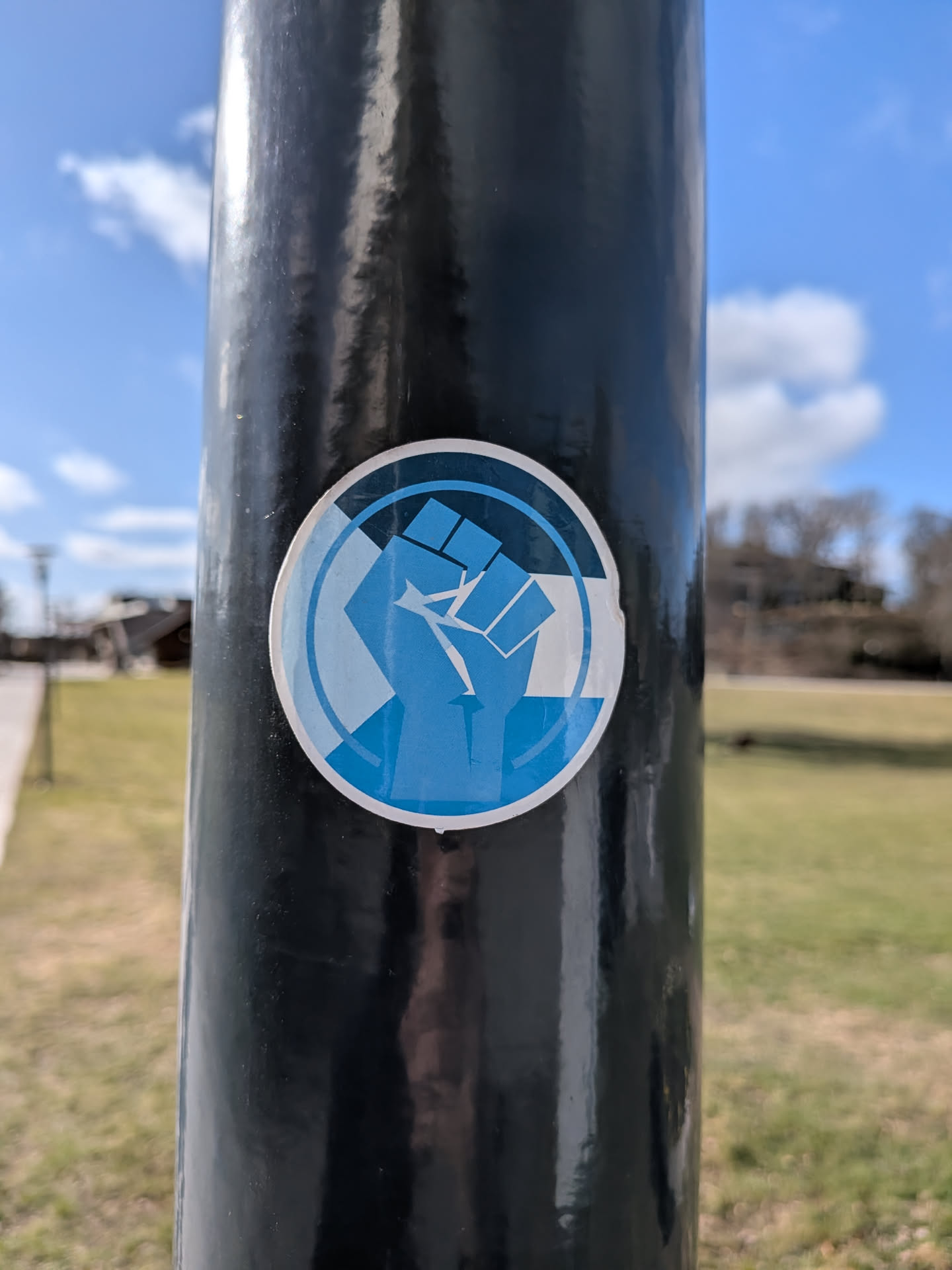 A circular sticker prominently featuring a stylized, upward-facing raised fist. The fist is depicted in a vibrant medium blue, composed of distinct geometric and somewhat pixelated segments. It is set against a background within the circle that is divided into sections of white and a lighter shade of blue, creating a dynamic visual effect. A darker blue outline encircles the entire design, framing the central motif. The sticker has a clean, graphic style and shows minor wear consistent with being affixed to a pole.