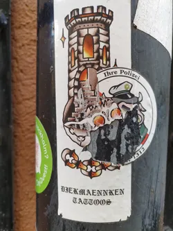 Street sticker A sticker from Djekmaenniken Tattoos. The main design is a traditional-style tattoo of a stone tower with a flame inside, and a small star above it. Below the tower is a partially damaged image of what appears to be a policeman's badge. The text on the badge reads "Ihre Polizei". The bottom of the sticker features the text "DJEKMAENNIKEN TATTOOS" in a Gothic-style font.