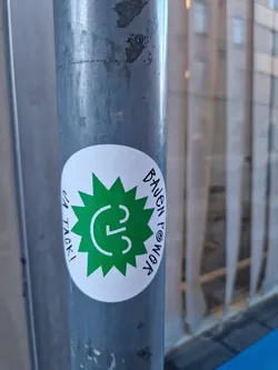 A round white sticker with a green sunburst design featuring the letter 'C' inside.  The text around the design says 'bauen powaga' and 'ein tacki!' in a handwritten style.  The sticker is affixed to a metal pole.
