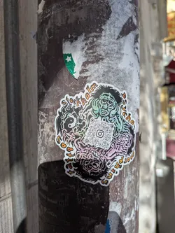 A die-cut sticker affixed to a weathered metal pole. The sticker features a central QR code containing the Instagram logo. Surrounding the QR code are colorful, psychedelic illustrations: a distressed green-skinned character holding its head at the top, a black and white yin-yang symbol on the left, and pink and purple brain-like textures at the bottom. The stylized text 'Stockholm Psycho Tattooing' is written in orange and black along the outer border.