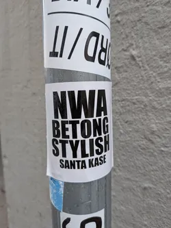 Street sticker A rectangular white sticker featuring bold, black, sans-serif text stacked vertically: 'NWA', 'BETONG', 'STYLISH', and 'SANTA KASE'. It is stuck to a metallic pole.