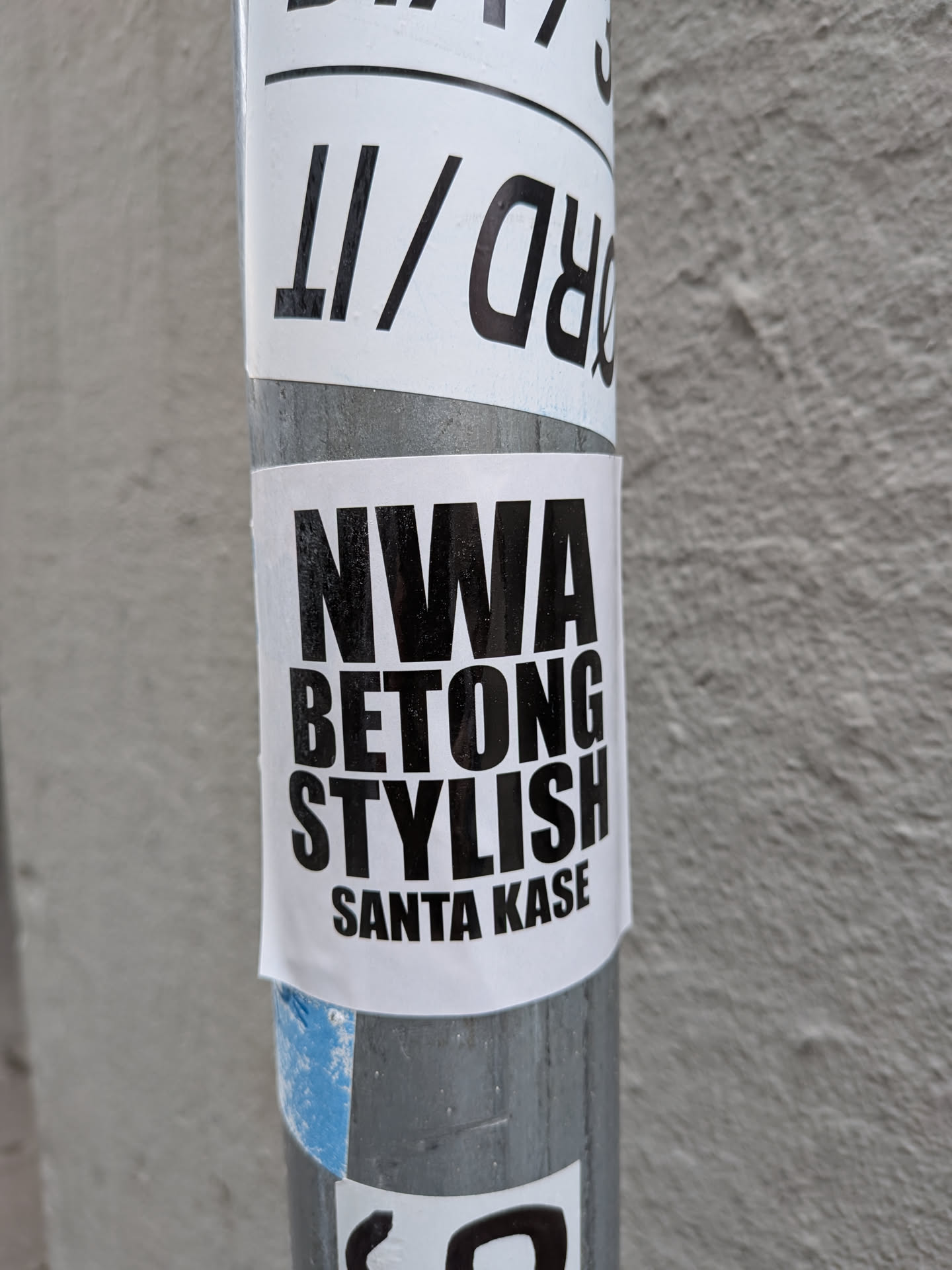 A rectangular white sticker featuring bold, black, sans-serif text stacked vertically: 'NWA', 'BETONG', 'STYLISH', and 'SANTA KASE'. It is stuck to a metallic pole.