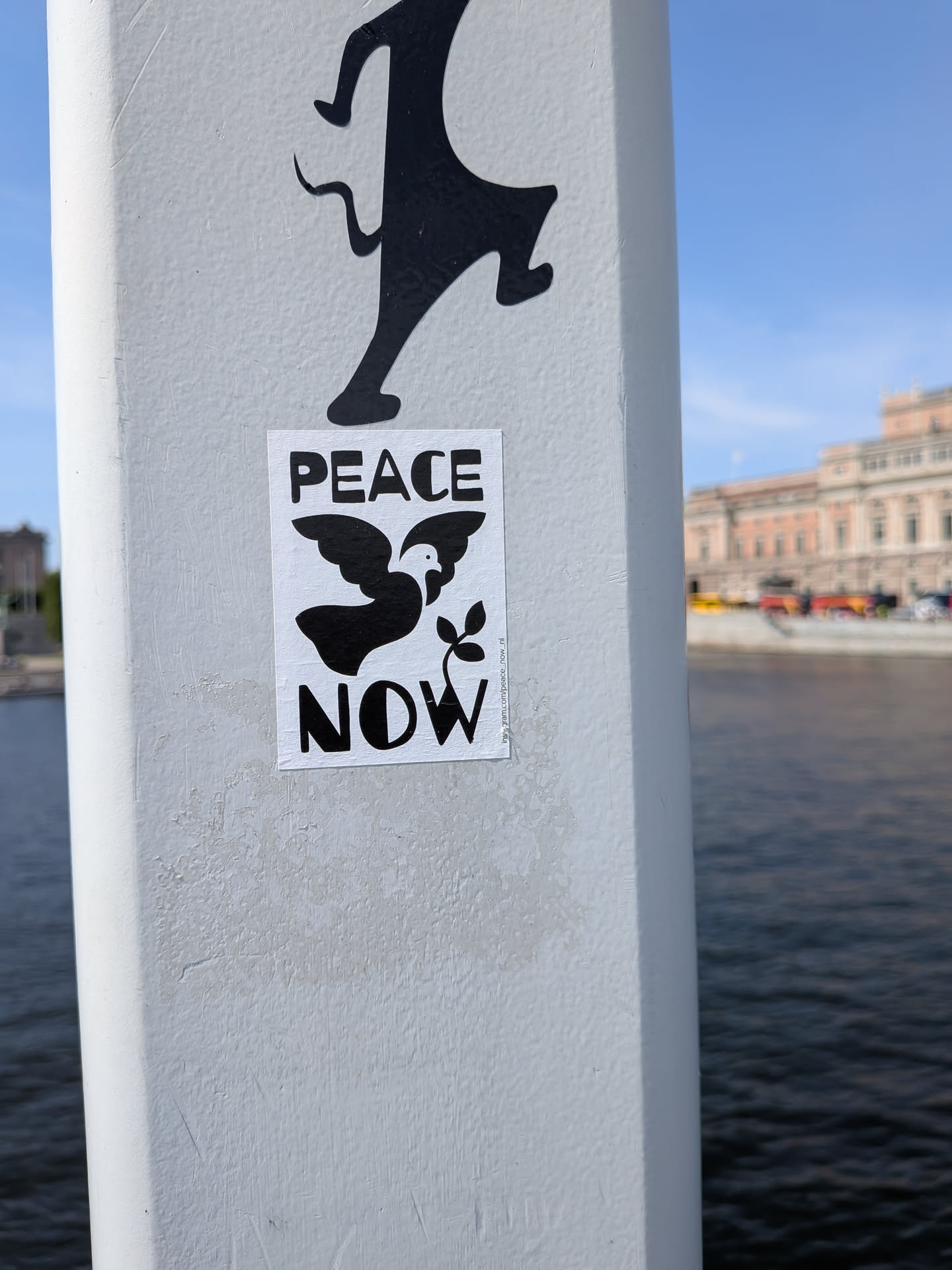A rectangular white sticker with bold black text reading 'PEACE' and 'NOW' framing a stylized black dove silhouette with an olive branch. A small Instagram handle is printed vertically on the right margin.