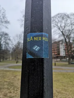 Street sticker A square-shaped sticker adhered to a dark, multi-faceted pole. The sticker features a dark blue background with a subtle texture resembling denim or a woven fabric. At the top, in bright yellow, sans-serif capital letters, is the Swedish phrase 'SLÅ NER MIG'. Below the text, centered, is a rectangular emblem with a dark border, containing two parallel light blue or white stripes on a darker blue field, strongly reminiscent of a military or police rank insignia. The sticker is slightly creased where it wraps around the pole's edges, indicating its physical presence and adherence.