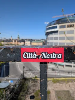 The sticker is a rectangular design with a vibrant red background. It prominently displays the text 'Città Nostra' in a bold, white, sans-serif font, outlined in black. Positioned between the words 'Città' and 'Nostra' is a stylized graphic of a fox head. The fox head features sharp, angular lines, primarily in red and black, with white details for the eyes and a small white, heart-shaped element beneath its chin. The overall aesthetic is clean, graphic, and impactful.