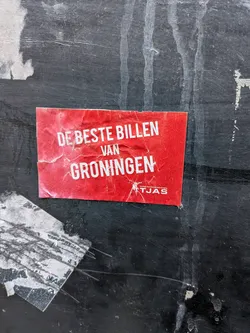 Street sticker A rectangular, vibrant red sticker adhered to a dark, weathered surface. The sticker is creased and slightly torn, showing signs of age and exposure. Prominently displayed in large, white, sans-serif capital letters is the phrase 'DE BESTE BILLEN VAN GRONINGEN' (The Best Butts of Groningen). Below this, in smaller white text, it reads 'Groninger Studenten Schaatsvereniging TJAS' (Groningen Student Skating Association TJAS), accompanied by a small white silhouette logo of a person ice skating. The overall appearance is that of a well-used, outdoor promotional sticker.