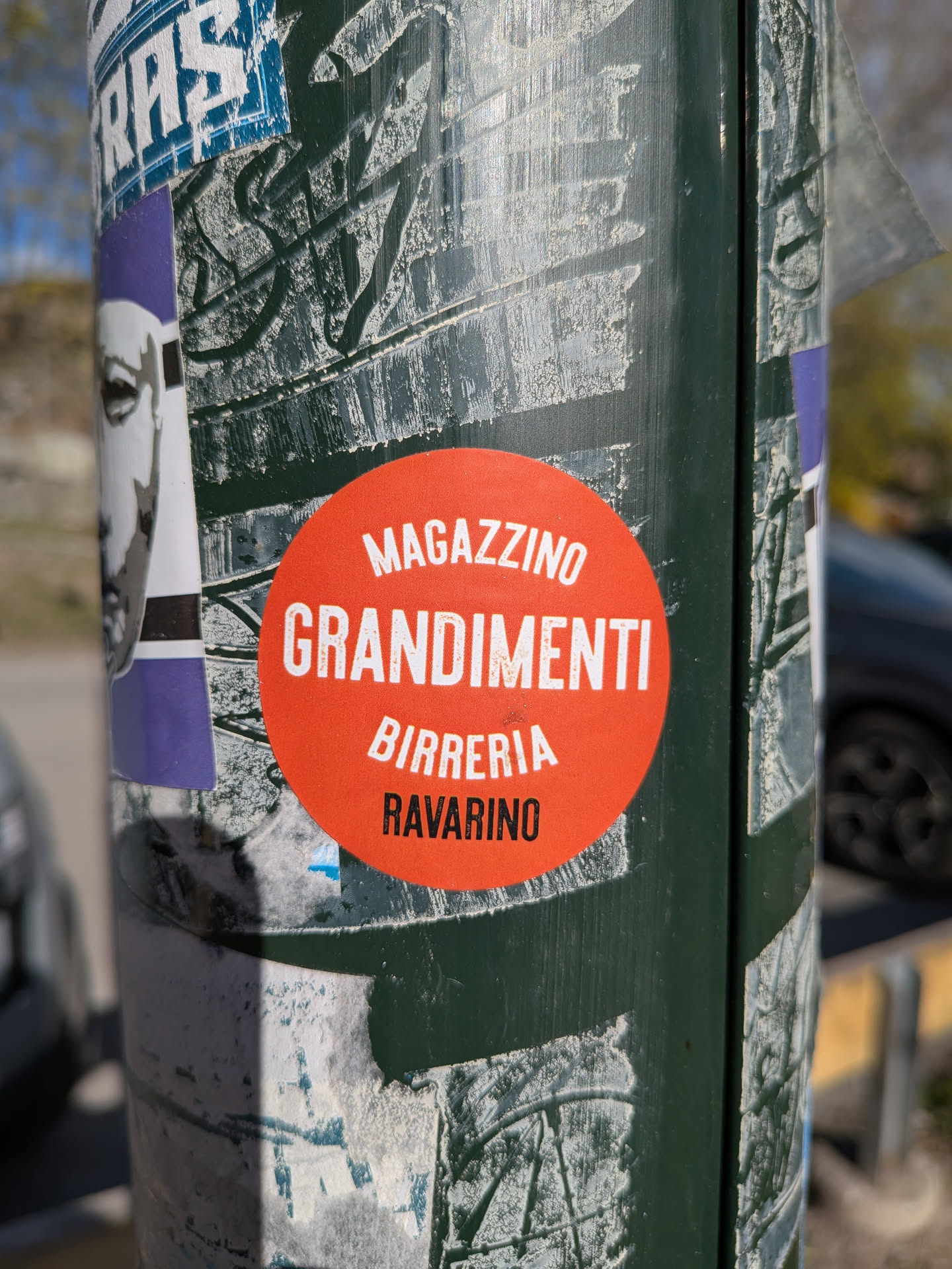 A vibrant, circular orange-red sticker prominently displayed on a dark green pole. The sticker features white, sans-serif, uppercase text arranged in four centered lines. The text reads 'MAGAZZINO', 'GRANDIMENTI', 'BIRRERIA', and 'RAVARINO'. The sticker appears relatively new and clean against the heavily weathered and stickered surface of the pole.