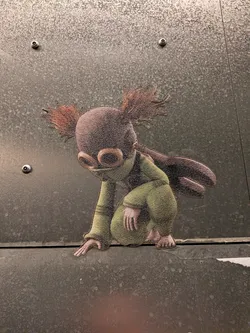 Street sticker The sticker depicts a small, humanoid creature with a rounded head, large, circular goggles, and spiky, reddish-brown hair that fans out. It is wearing a green, hooded garment that covers its torso and legs. The creature is in a crouching or kneeling position, with one hand extended forward and the other resting on its knee. Its feet are visible and appear to have toes. The overall color palette is muted, with earthy tones and greens, and the sticker has a textured appearance.