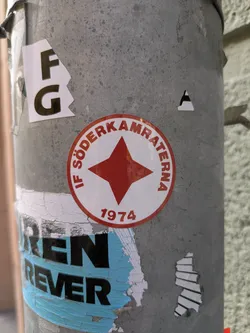 Street sticker A round sticker affixed to a metal pole. The sticker is white with a red border and a red five-pointed star in the center. The text "IF SÖDERKAMRATERNA" is printed in a semi-circle above the star, and the number "1974" is printed below the star. The sticker appears to be old and slightly faded. There are other stickers and remnants of stickers on the pole.