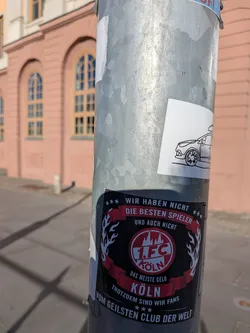 Street sticker The sticker is predominantly black with red and white accents. At the center is a circular emblem featuring '1. FC KÖLN' in white text within a red circle, surrounded by a white outline and a stylized mountain graphic. Above the emblem, a red banner reads 'WIR HABEN NICHT DIE BESTEN SPIELER UND AUCH NICHT'. Below the emblem, another red banner states 'DAS MEISTE GELD KÖLN'. The bottom of the sticker features text that reads 'TROTZDEM SIND WIR FANS VOM GEILSTEN CLUB DER WELT'. The emblem is flanked by stylized white flames or wings. There is also a smaller, separate sticker above it featuring a minimalist black outline drawing of a car.