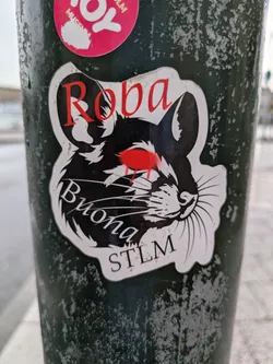 Street sticker The sticker is predominantly black and white with striking red accents, depicting the head of a rodent, likely a mouse or rat, in profile, facing right. The rodent has large, pointed ears, prominent whiskers, and an expressive, almost fierce, look. Its eyes are a vivid, dripping red, giving it an intense appearance. The fur is rendered with jagged, sharp lines, creating a somewhat aggressive or stylized texture. Above the rodent's head, the word "Roba" is written in red, stylized script. Below the head, "Buona" is written in white, cursive script, and "STLM" is in white, block letters. The sticker has a distinct white outline, giving it a cut-out appearance against the dark, textured pole it's affixed to.