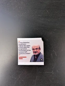 Street sticker A square-shaped sticker featuring a quote by Salman Rushdie on a white background. The left side displays the quote in dark grey text, framed by large quotation marks. The right side contains a color portrait of Salman Rushdie, an older man with glasses, a white beard, and a slight smile, wearing a dark shirt and grey blazer. Below the quote, in small orange-red boxes, are the words 'SALMAN RUSHDIE' and 'WRITER'. A small, dark, stylized 'M' logo is visible in the bottom right corner of the portrait. The sticker has a slightly torn edge on the top right.