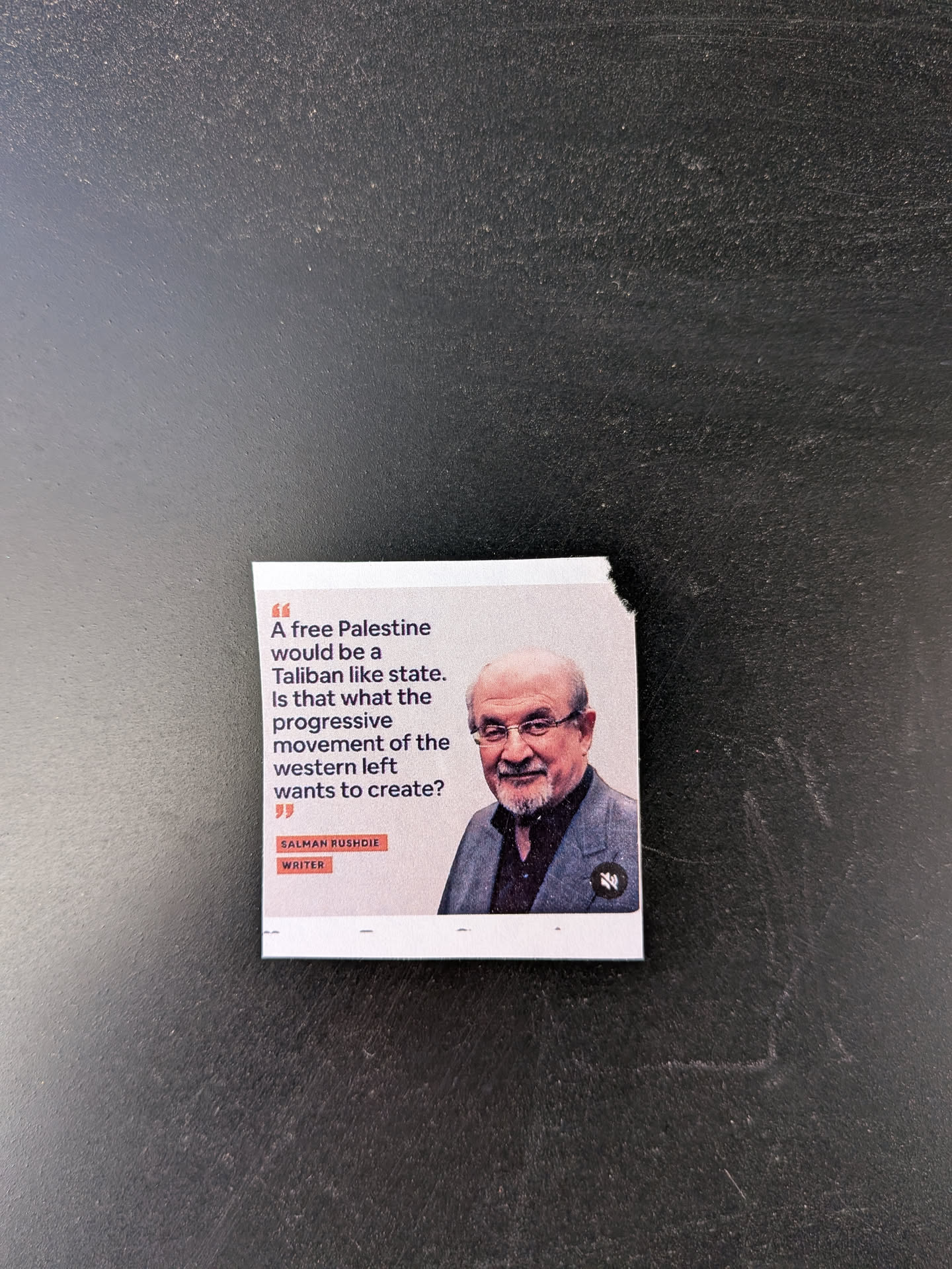 A square-shaped sticker featuring a quote by Salman Rushdie on a white background. The left side displays the quote in dark grey text, framed by large quotation marks. The right side contains a color portrait of Salman Rushdie, an older man with glasses, a white beard, and a slight smile, wearing a dark shirt and grey blazer. Below the quote, in small orange-red boxes, are the words 'SALMAN RUSHDIE' and 'WRITER'. A small, dark, stylized 'M' logo is visible in the bottom right corner of the portrait. The sticker has a slightly torn edge on the top right.