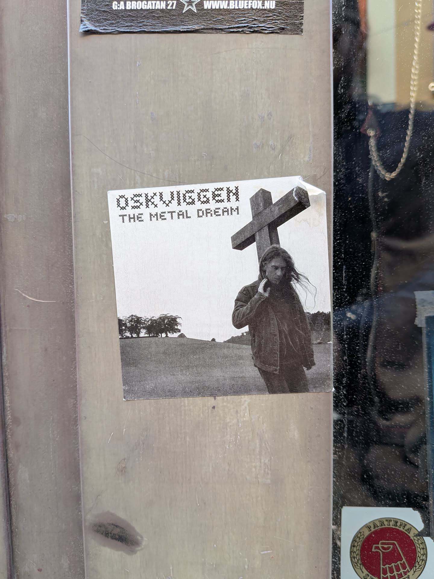 Square black-and-white sticker showing a long-haired figure carrying a large wooden cross across a field. Stylized cross-stitch text in the upper left reads 'OSKVIGGEN' and 'THE METAL DREAM'.