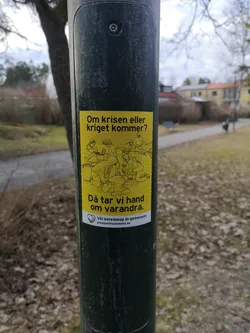 Street sticker The sticker is rectangular with a vibrant yellow background and features black line art illustrations. At the top, the text "Om krisen eller kriget kommer?" is written. Below this, a group of approximately seven stylized figures are depicted in various acts of assistance and preparation, including tending to an injured person, cooking, and gathering resources like firewood. Below the illustration, the text "Då tar vi hand om varandra." is displayed. At the bottom, a small white heart icon containing two shaking hands is present, followed by the text "Vår beredskap är gemensam" and the website address "preppatillsammans.se". The overall aesthetic is simple, illustrative, and conveys a strong message of collective action and support.