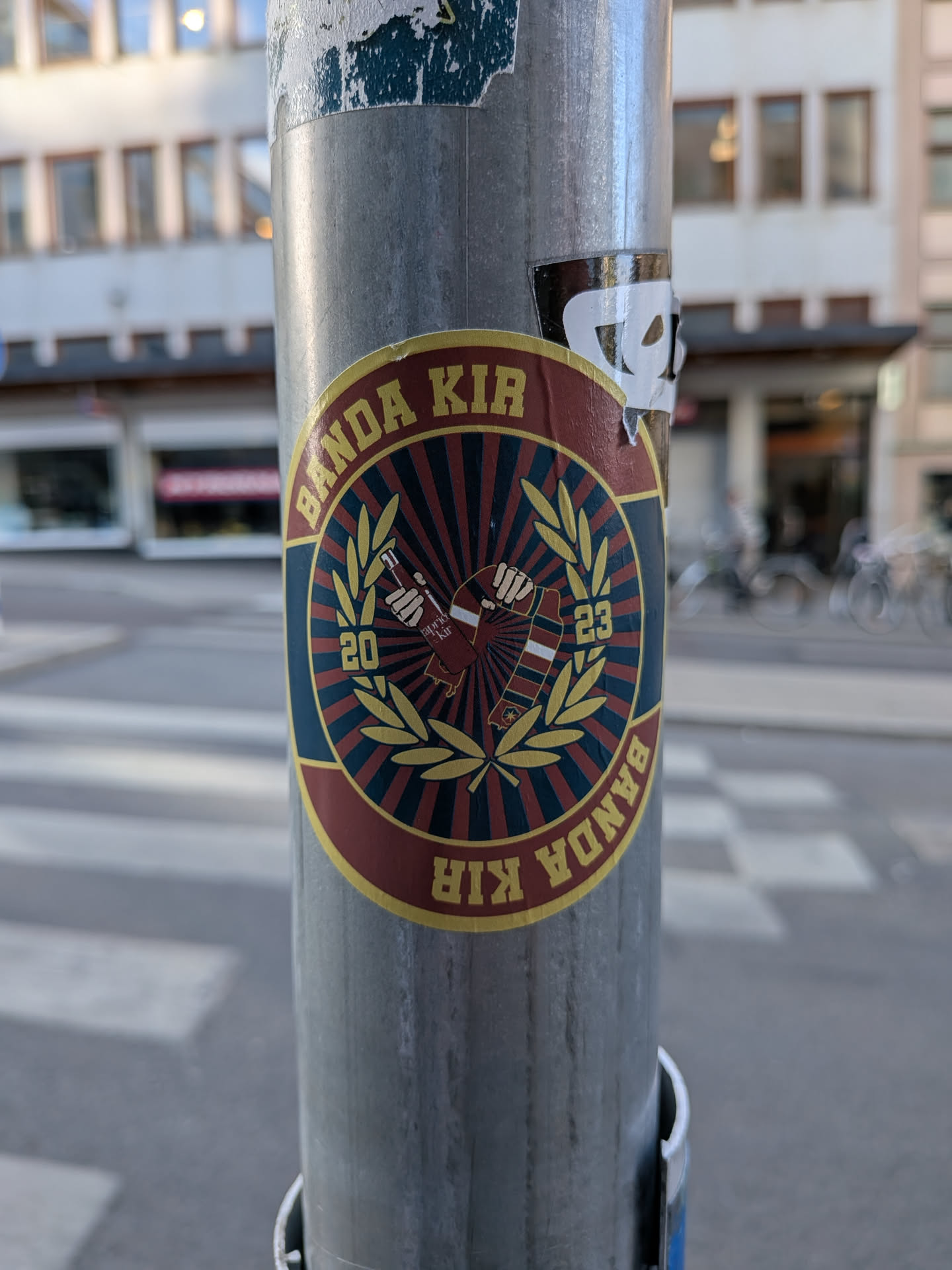 A round sticker featuring a red and blue sunburst background enclosed by a golden laurel wreath. Two hands in the center hold a bottle and a striped scarf. The outer maroon border displays the yellow text 'BANDA KIR' and the year '2023'.