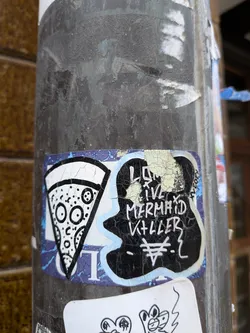 Street sticker The sticker is applied to a dark, textured pole and shows significant wear and tear, with peeling edges and cracked surfaces. It has a blue border that is partially visible and faded. The main design is split into two distinct parts. On the left, there is a stylized, black and white illustration of a pizza slice. The crust is white, and the body of the pizza is white with various black circular shapes representing toppings, some of which resemble eyes and a mouth, giving the pizza a cartoonish, somewhat expressive face. On the right, there is an irregular, black blob-like shape with white, hand-drawn text that reads 'LONG LIVE MERMAID KILLER'. Below this text, there is a unique white symbol resembling a stylized '¥' or a trident with a horizontal bar. The black area of this section is heavily cracked, revealing lighter tones underneath. A small, vertical 'K2021' is visible on the far right edge of the black blob.