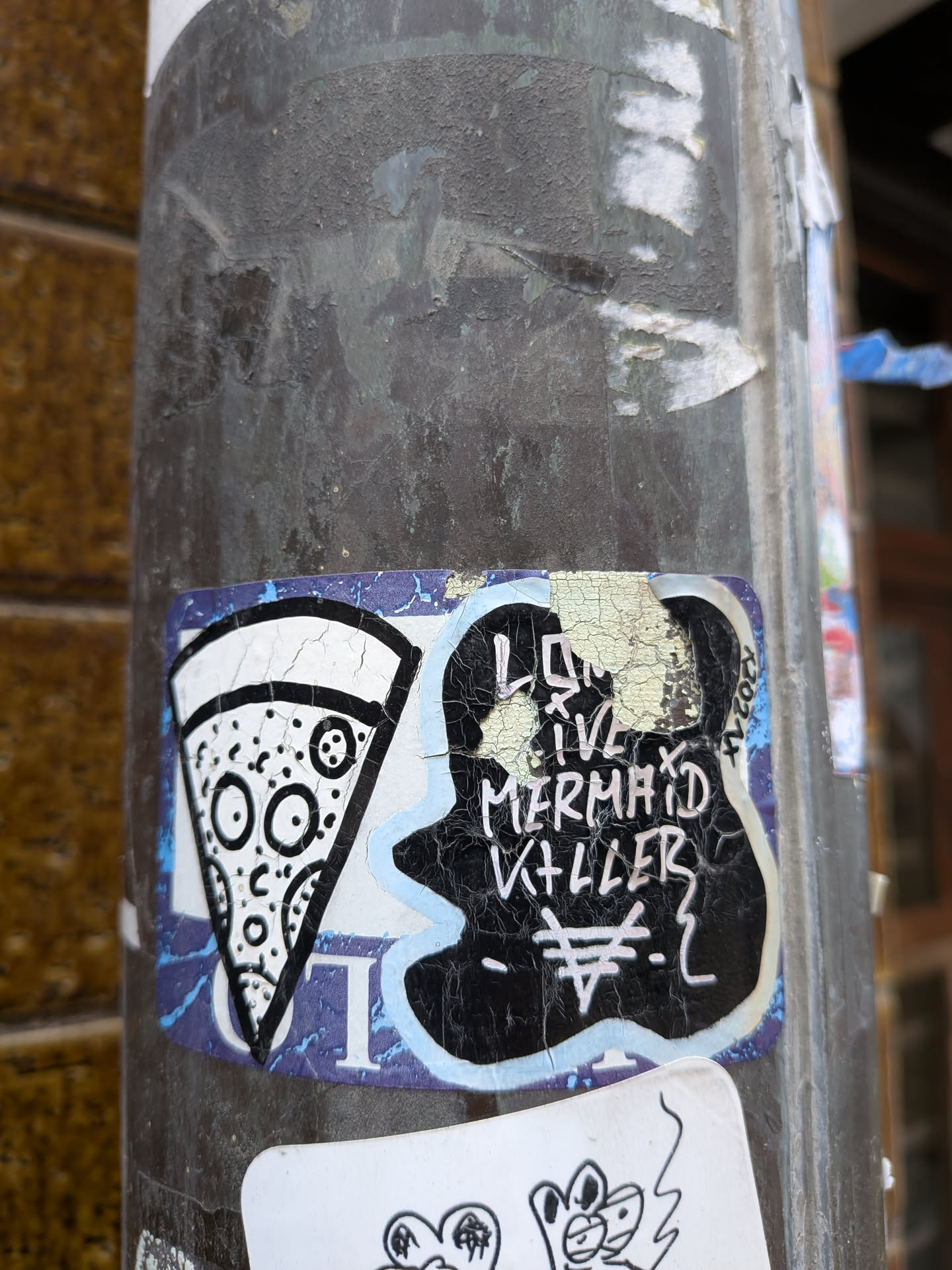 The sticker is applied to a dark, textured pole and shows significant wear and tear, with peeling edges and cracked surfaces. It has a blue border that is partially visible and faded. The main design is split into two distinct parts. On the left, there is a stylized, black and white illustration of a pizza slice. The crust is white, and the body of the pizza is white with various black circular shapes representing toppings, some of which resemble eyes and a mouth, giving the pizza a cartoonish, somewhat expressive face. On the right, there is an irregular, black blob-like shape with white, hand-drawn text that reads 'LONG LIVE MERMAID KILLER'. Below this text, there is a unique white symbol resembling a stylized '¥' or a trident with a horizontal bar. The black area of this section is heavily cracked, revealing lighter tones underneath. A small, vertical 'K2021' is visible on the far right edge of the black blob.