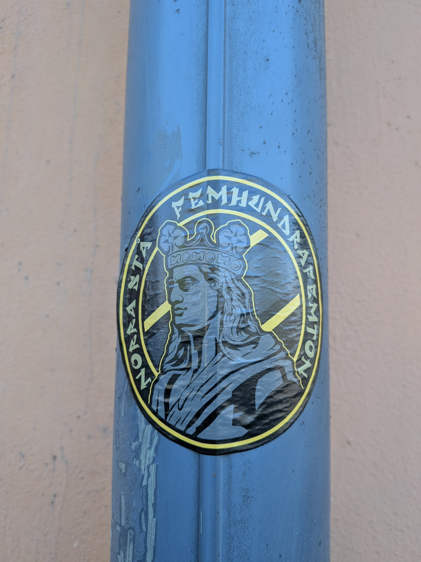 The sticker is circular, predominantly black and yellow, with a central profile portrait of a person wearing an ornate crown. The figure is rendered in shades of grey and black, with a strong, illustrative style. The word 'MOMON' is visible on the front of the crown. The background behind the figure consists of diagonal yellow stripes on a black field. A yellow border encircles the central image, containing black text. The text reads 'FEMHUNDRA LAPPAR' across the top arc and 'NORRASTA' along the bottom left arc. The sticker has a slightly worn appearance with minor creases, indicating it's a physical sticker applied to a surface.