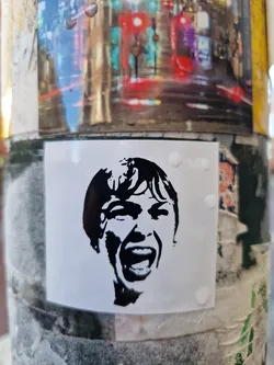 Black and white stencil art of a woman screaming. The sticker is affixed to a pole covered in other stickers and posters.
