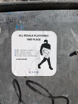 Street sticker A white, rounded-rectangle sticker adhered to a dark, textured metal surface. The sticker features a minimalist design with black text and a black silhouette graphic. At the top, bold black text reads 'ALL RESALE PLATFORMS' and 'ONE PLACE'. Below this, a list of bullet points in blue squares details features: '100+ Designer brands', '500+ Resale stores', 'Customizable filters', and 'Instant drop notifications'. To the right, a black silhouette of a person, possibly wearing a balaclava, walks left while carrying a large rectangular screen displaying 'BUY NOW' and a designer-style handbag. A thin white line extends from the person's hand to a small blue oval logo at the bottom center, which contains 'K • U • R • B' in white text. The website 'KURB.ONLINE' is printed above this logo on the left. The sticker shows minor creases and dirt, indicating it's a physical object.