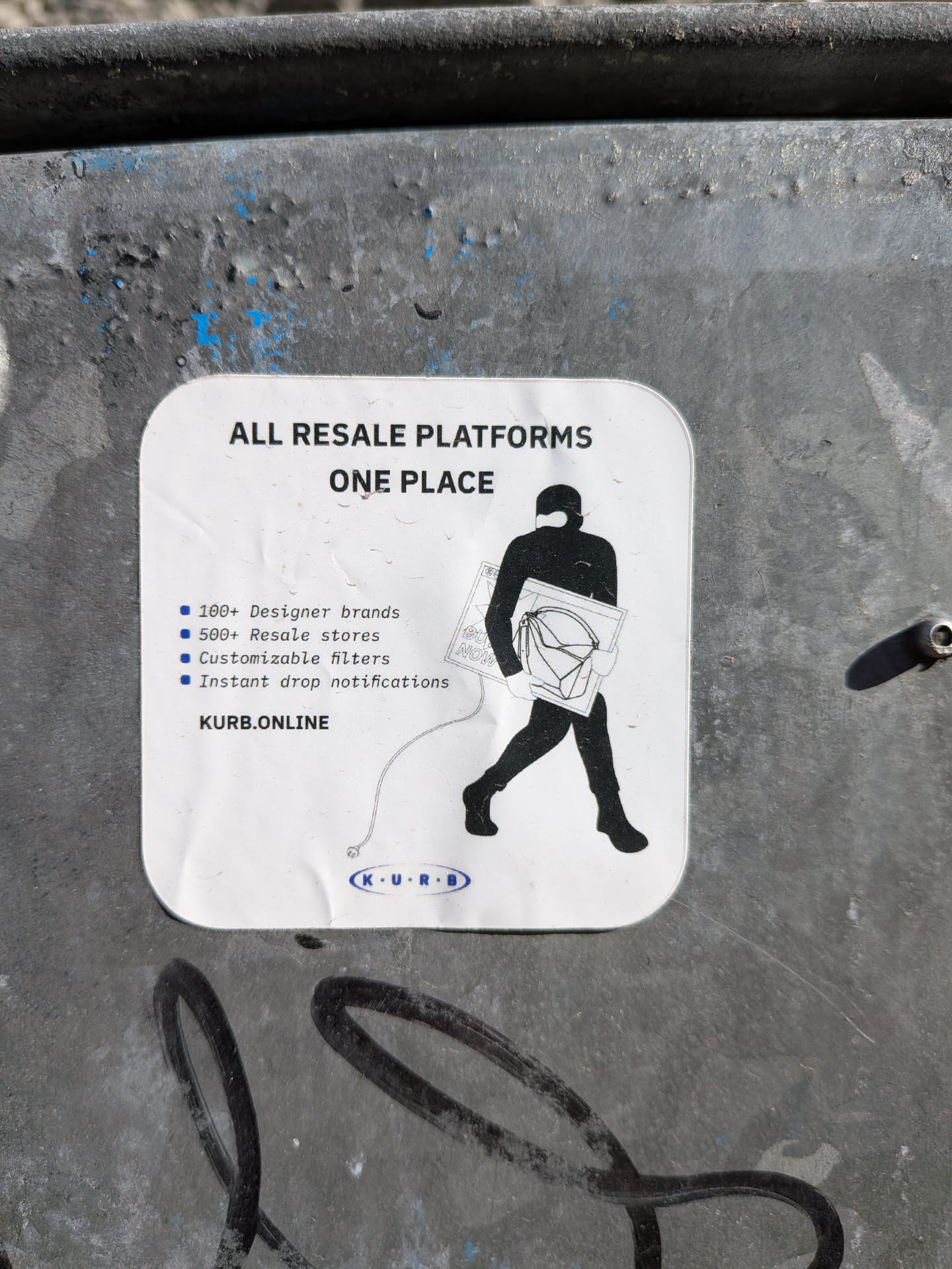 A white, rounded-rectangle sticker adhered to a dark, textured metal surface. The sticker features a minimalist design with black text and a black silhouette graphic. At the top, bold black text reads 'ALL RESALE PLATFORMS' and 'ONE PLACE'. Below this, a list of bullet points in blue squares details features: '100+ Designer brands', '500+ Resale stores', 'Customizable filters', and 'Instant drop notifications'. To the right, a black silhouette of a person, possibly wearing a balaclava, walks left while carrying a large rectangular screen displaying 'BUY NOW' and a designer-style handbag. A thin white line extends from the person's hand to a small blue oval logo at the bottom center, which contains 'K • U • R • B' in white text. The website 'KURB.ONLINE' is printed above this logo on the left. The sticker shows minor creases and dirt, indicating it's a physical object.