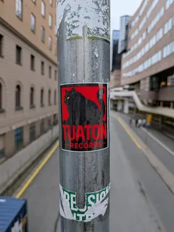Street sticker The sticker features a silhouette of a black cat with bright yellow eyes, positioned centrally against a solid red background. The cat is depicted in a stylized manner, with its tail curved upwards. Below the cat, the text 'TUATON RECORDS' is prominently displayed in bold, red, sans-serif capital letters, with 'RECORDS' underlined by a thin red line. The sticker has a white border and appears to be slightly worn or creased, indicating it has been affixed to the pole for some time.
