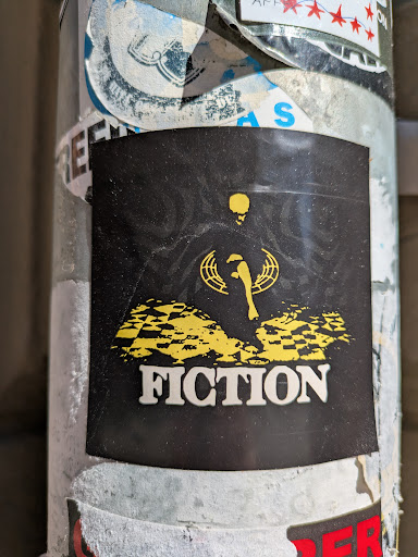 The sticker has a black background with a yellow figure above the word "FICTION". The figure appears to be a person or skeleton holding a scepter, surrounded by a geometric design. The word "FICTION" is printed in a bold, white font.