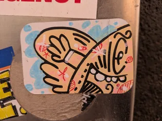 Street sticker A cartoon face sticker with a light tan base color and black outlines. The face is framed by two hands in a similar style. The background has light blue accents resembling water droplets. The artwork uses pink and red for additional accents. The face features exaggerated features such as a large eye, prominent teeth, and other whimsical details.