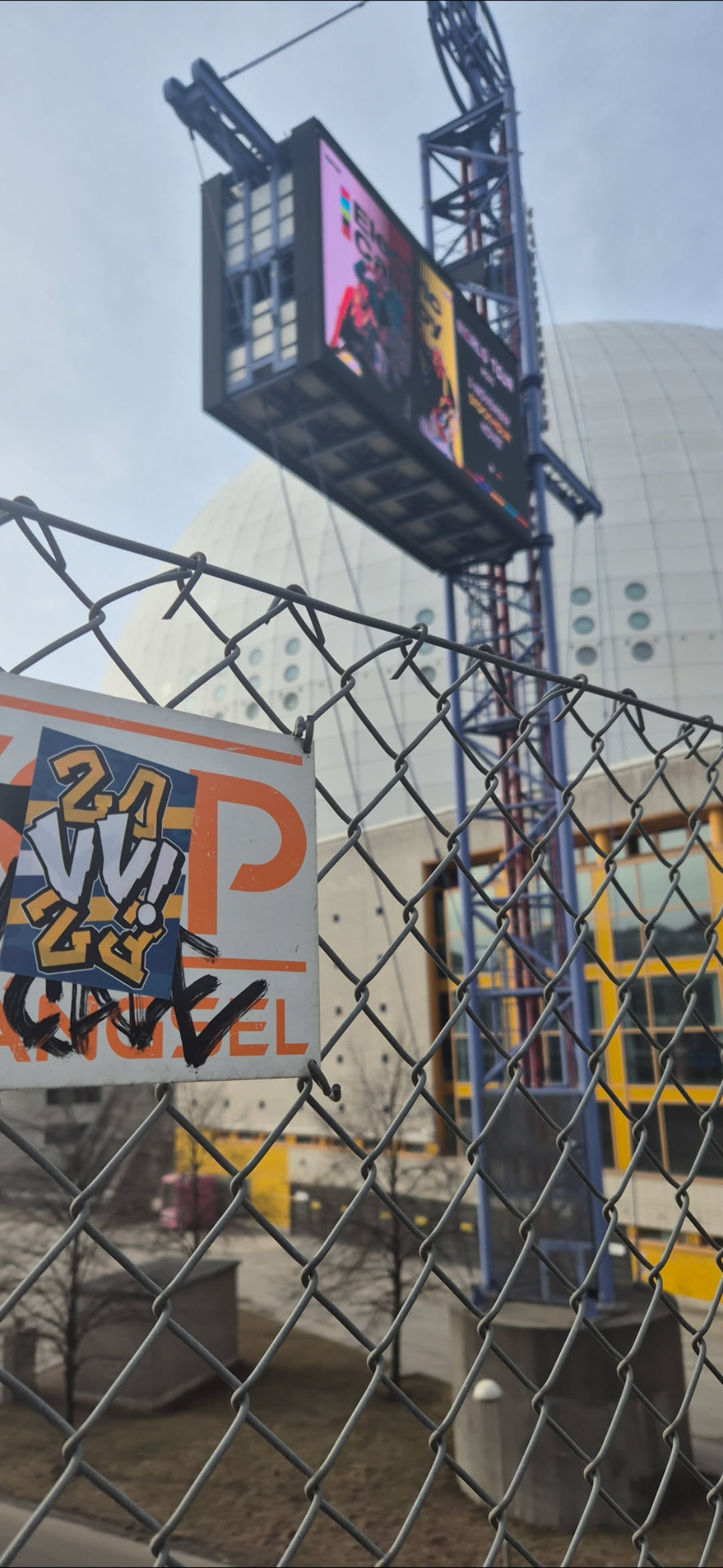 A close-up shot captures a rectangular sticker affixed to a chain-link fence. The sticker features the text "24 VI 2023" in bold letters and the name "GRRRL" written beneath. The colors of the text and background of the sticker are a mix of orange, white, and blue. The fence is a gray chain-link construction. The backdrop features a large, spherical building under a cloudy sky.