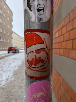 The sticker features a stylized, hand-drawn depiction of what appears to be a Santa hat in vibrant orange and red tones, with a white brim and pom-pom. Below the hat, there's a round, white shape with irregular red/orange splotches, possibly representing a face or body, outlined in black. The overall style is cartoonish and somewhat abstract, with a distinct, slightly rough black outline defining the elements. Water droplets are visible on its surface, indicating it's exposed to the elements.