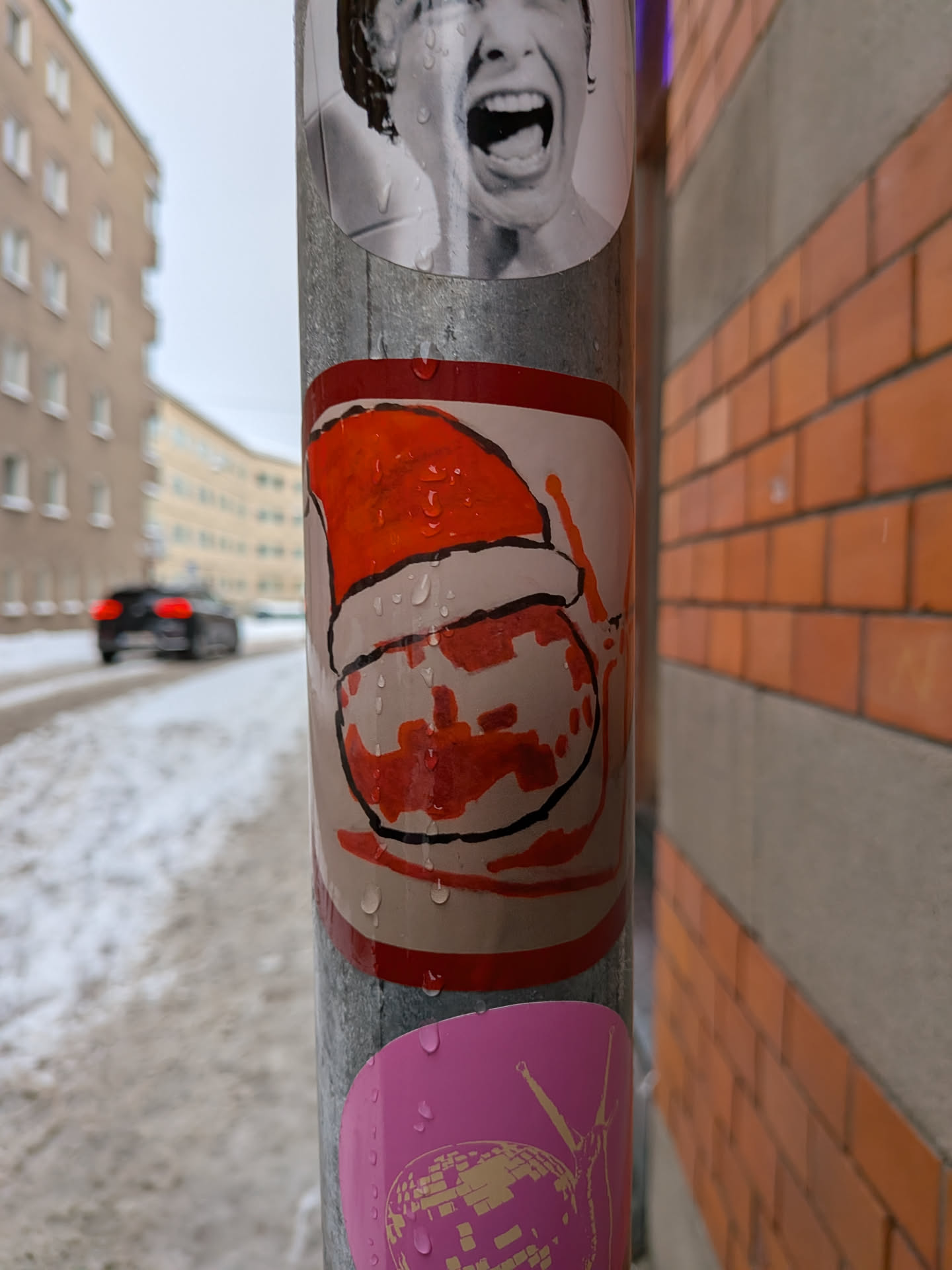 Street sticker The sticker features a stylized, hand-drawn depiction of what appears to be a Santa hat in vibrant orange and red tones, with a white brim and pom-pom. Below the hat, there's a round, white shape with irregular red/orange splotches, possibly representing a face or body, outlined in black. The overall style is cartoonish and somewhat abstract, with a distinct, slightly rough black outline defining the elements. Water droplets are visible on its surface, indicating it's exposed to the elements.