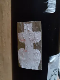 Close-up of a small, white sticker affixed to a dark-colored pole. The sticker features a minimalist design with a somewhat abstract central image. The sticker shows signs of wrinkling and discoloration, possibly from environmental exposure.